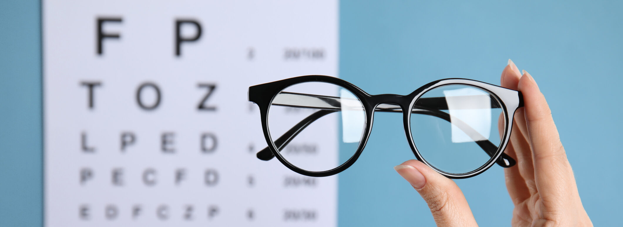 A hand holds a pair of prescription eyeglasses in front of a Snellen chart.